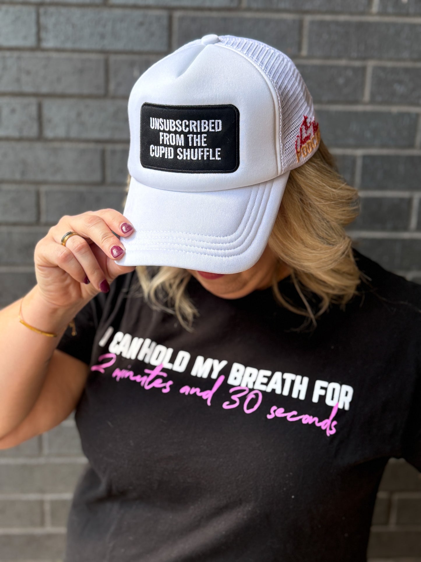Person wearing a white cap with text and a black t-shirt with humorous text against a brick wall.